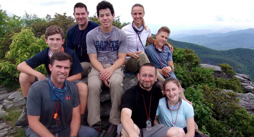 A group of parents and children pose for a photo on an overlook. There is a mountainous landscape in the background.